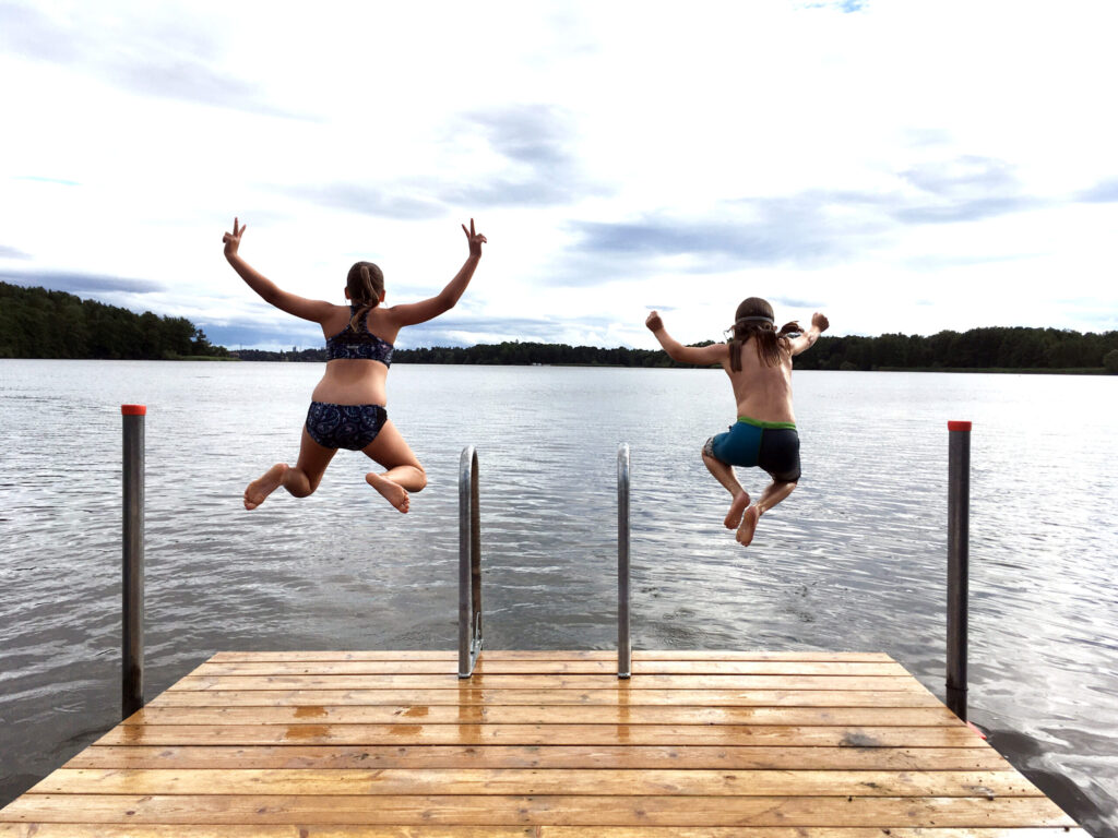 Två barn i badkläder som hoppar från en brygga ned i vattnet.
