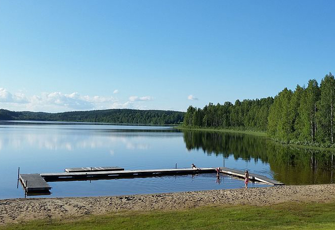 En badplats med stilla vatten, skog och en blå himmel.