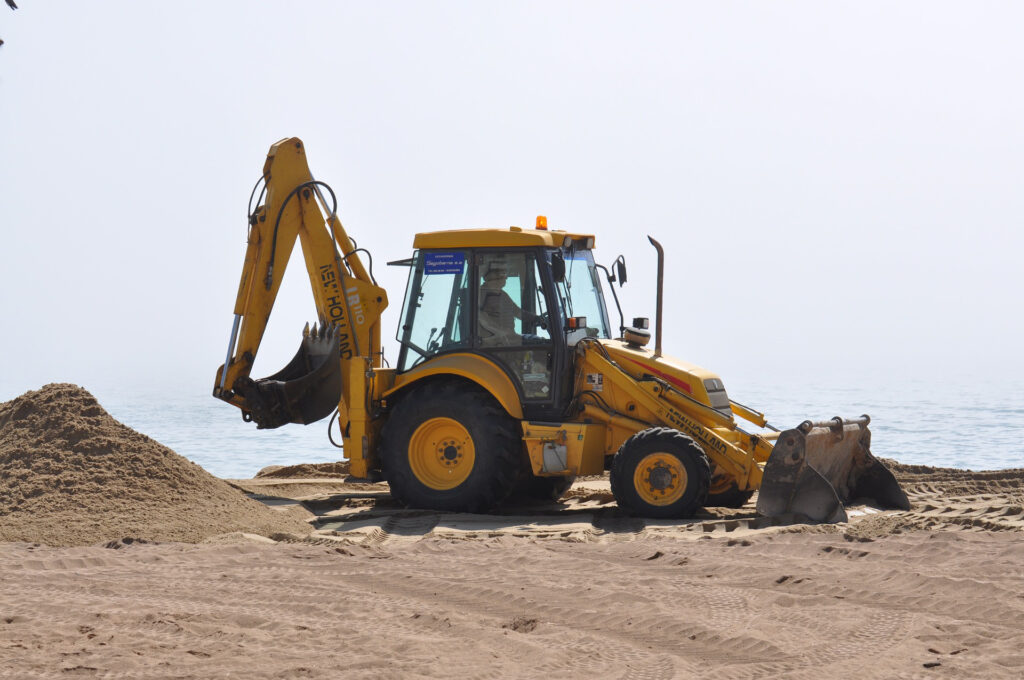 En orange grävskopa på en sandstrand med vatten i bakgrunden.