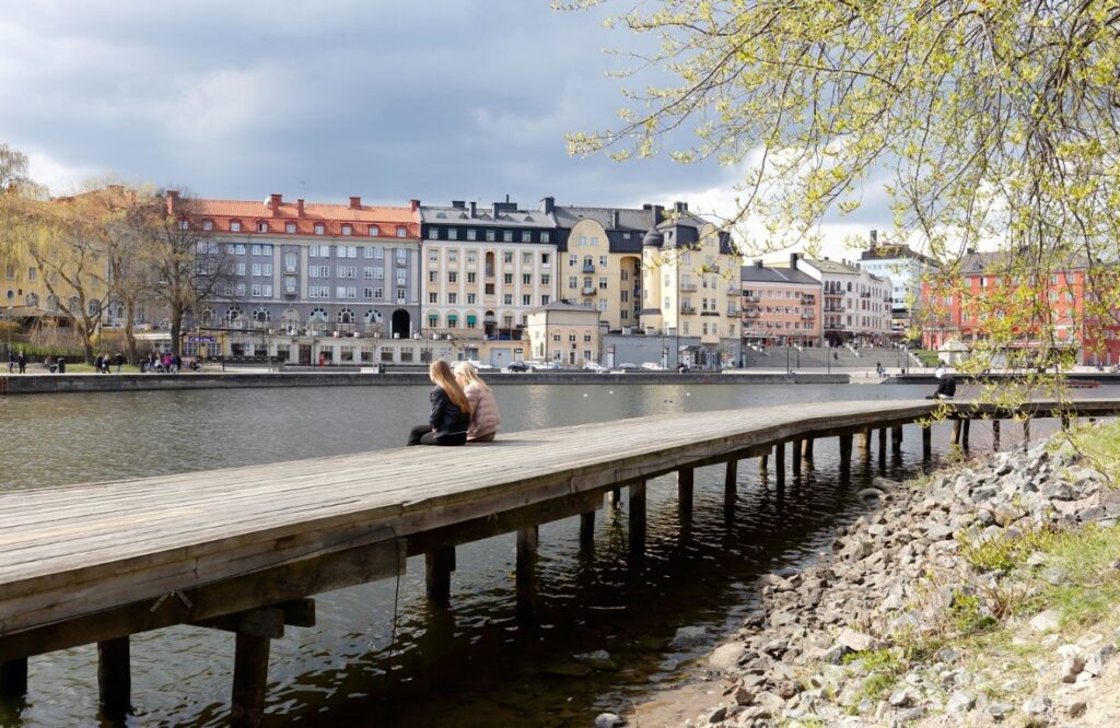 Två tjejer sitter på en lång träbrygga vid ett vattendrag i en stor stad.