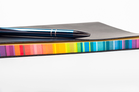 Ett svart anteckningsblock med färgade sidor och en penna.